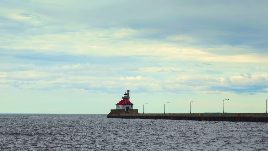 Historic Duluth South Breakwater Outer Light stands on concrete pier extending into Lake Superior, white building with red hip roof and green lantern room under dramatic cloudy sky.