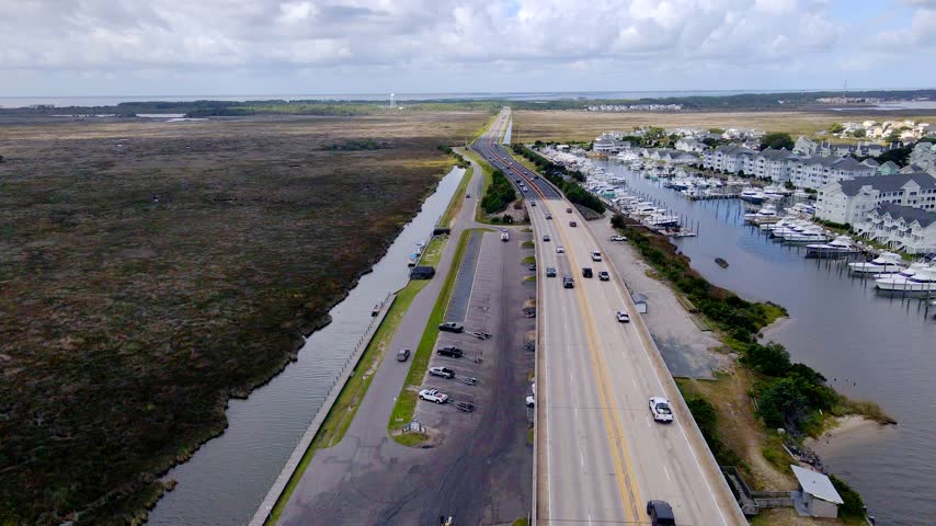 Cars travel along a coastal road between broad marshland and the marina waterfront.