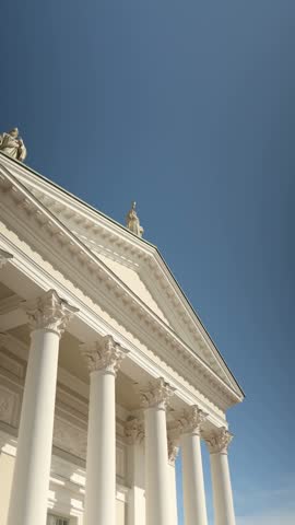 Vertical screen low angle view of Helsinki Cathedral facade with tall white columns triangular pediment rooftop statues and dome against clear blue sky in sunny Finland