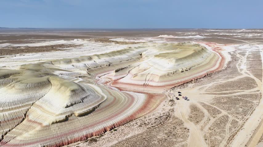 A close aerial orbit moves around two rounded chalk formations wrapped in red, pink and gray layers, with desert tracks and small visitors visible below.