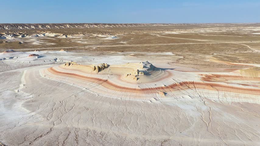 An aerial pass frames an isolated cream-colored butte enclosed by curved red bands and deeply scored white slopes in the Kyzylkup badlands.