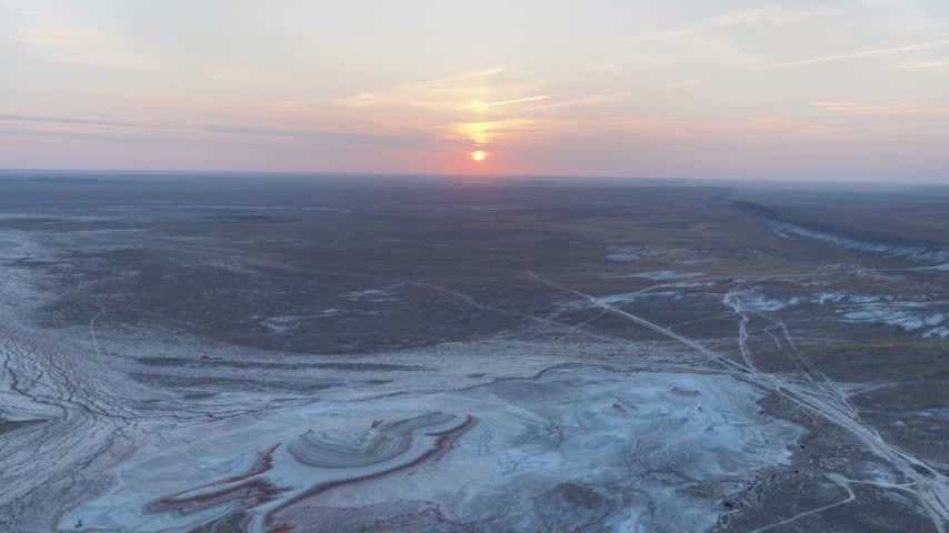 The orange sun hangs above the horizon as a high drone view drifts across pale drainage channels, dark plains and a small red-ringed hill at Kyzylkup.