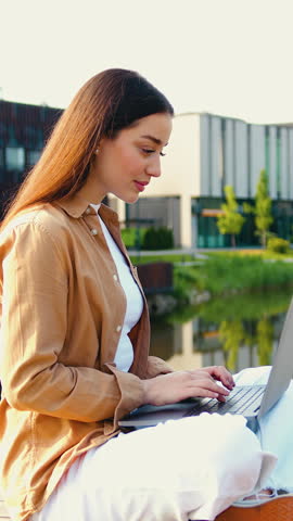 Concentrated young businesswoman using a laptop, wearing an earpiece, sitting cross-legged in a park near a lake, working remotely in a calm and natural setting. Slow motion
