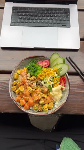 Vertical static top down view of a salmon poke bowl with avocado in front of an open laptop on an outdoor wooden table in early afternoon daylight, Berlin, Germany