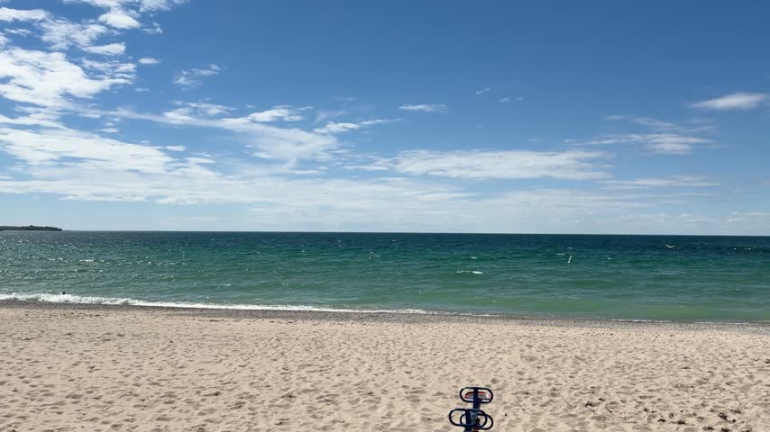 Peaceful view of Lake Michigan in Charlevoix, Michigan, with expansive blue sky, scattered white clouds and gentle waves along the sandy shoreline on a summer day.
