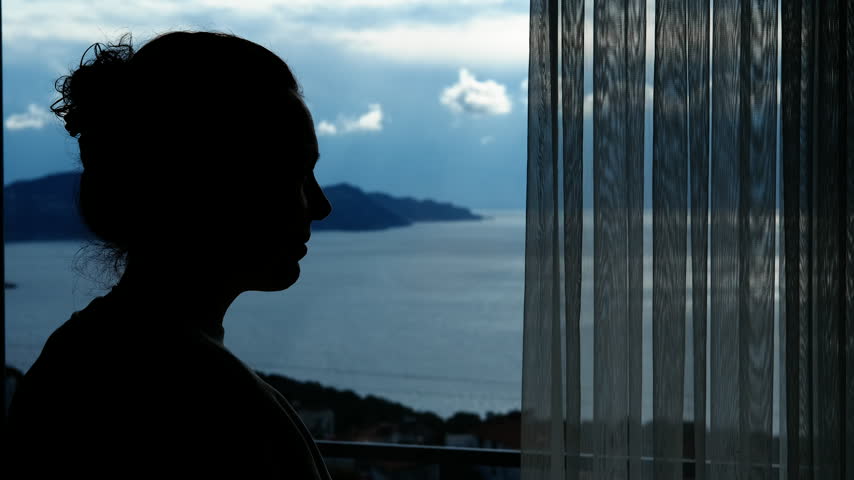 Silhouette of a pensive woman looking out window at sea. Young thoughtful woman standing in silhouette at a window, pensively gazing at the serene sea and islands landscape outside
