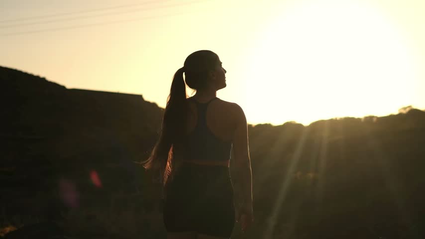 Woman celebrates victory with arms raised toward a mountain sunset, forming a V shape in golden light. Her silhouette and smartwatch express outdoor fitness, achievement, wellness, confidence.