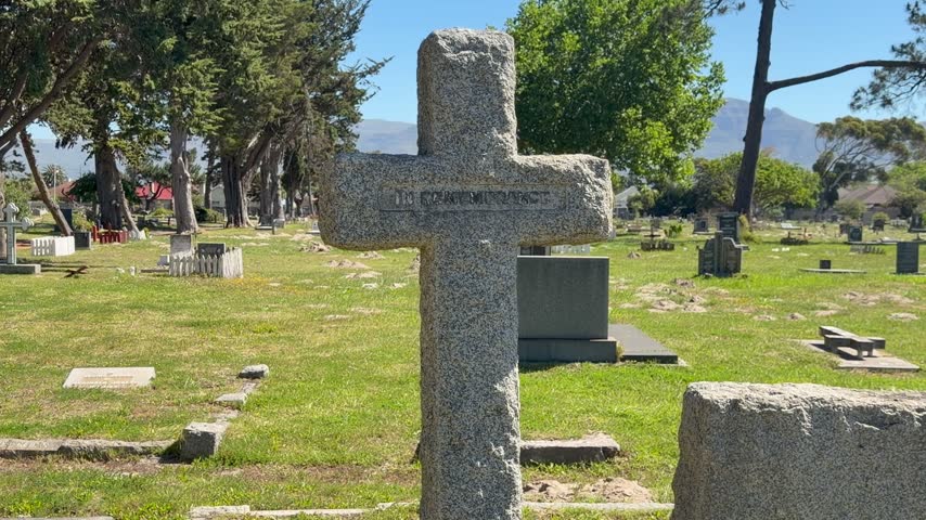 Close-up of a weathered granite cross grave marker in a peaceful cemetery on a sunny day, with green grass, tall trees and distant mountains under a clear blue sky.