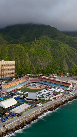 Cinematic aerial shot of a colorful baseball stadium repurposed with white tents, located between a lush green mountain and the turquoise ocean. Ideal for humanitarian or event logistics themes.