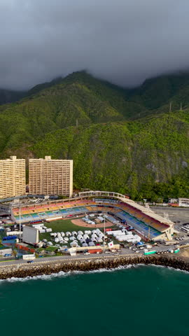 Cinematic aerial drone shot of a colorful baseball stadium used as a field hospital or emergency shelter, located between the ocean and lush green mountains.