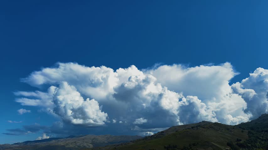 huge, billowing white cumulus clouds accumulating rapidly over a rolling mountain peak under a bright blue sky.