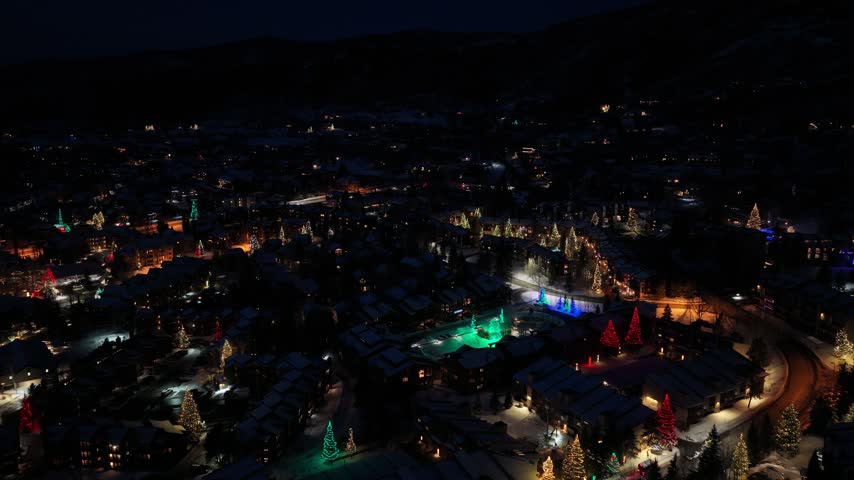 Winter drone footage of Whitefish, Montana, captures a snow covered mountain town at night with glowing streets, festive Christmas trees, lodges, and dark hills beyond the village.