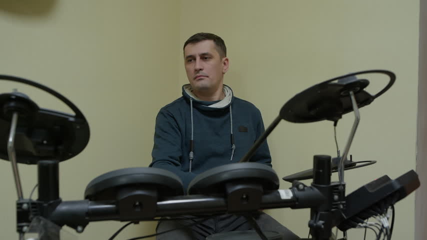 Man wearing blue hoodie sitting behind electronic drum kit, playing with drumsticks, focused expression, yellow wall background, black drum pads, practice session indoors