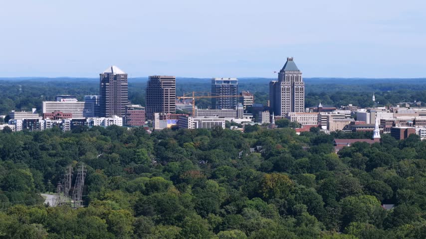 Telephoto view of Greensboro downtown with a Marriott hotel wordmark, a pyramid topped tower, a construction crane and power pylons above the treetops in Greensboro, North Carolina, USA. Aerial view