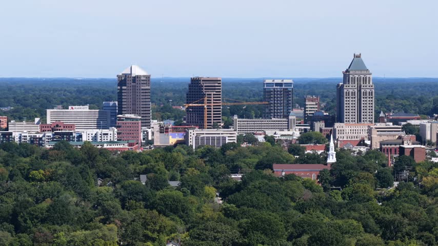 Telephoto view of Greensboro towers behind a green canopy, a Marriott hotel wordmark on a low building, a pyramid topped tower and a white steeple, in Greensboro, North Carolina, USA. Aerial view