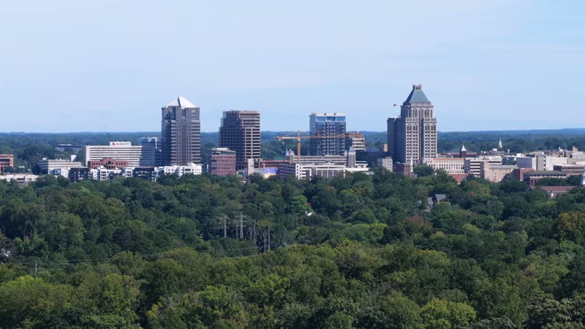 Greensboro skyline with a Marriott hotel wordmark, a pyramid topped tower and a crane rises above a wide band of green trees with power pylons in Greensboro, North Carolina, USA. Aerial view