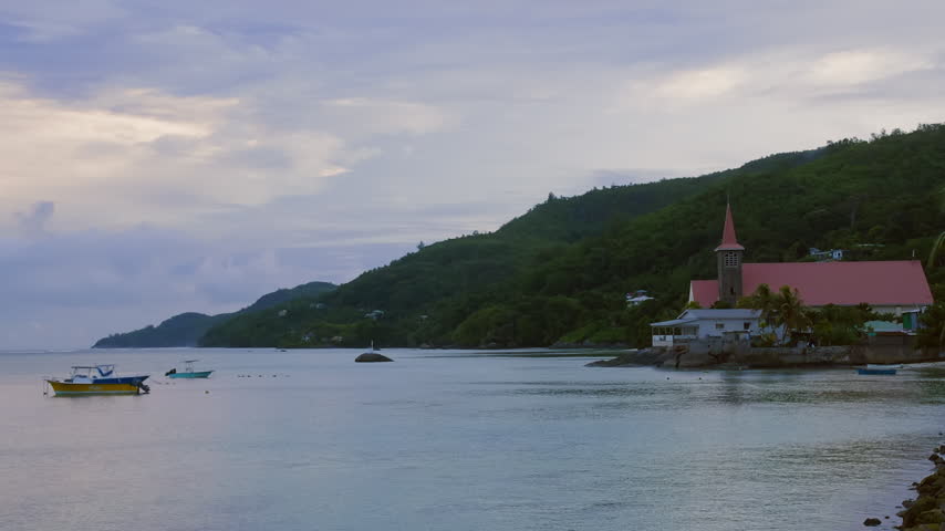 Seaside chapel featuring pink roof perched on rocky peninsula, fishing boats anchoring in shallow bay, lush forested mountains rising behind, overcast tropical sky.