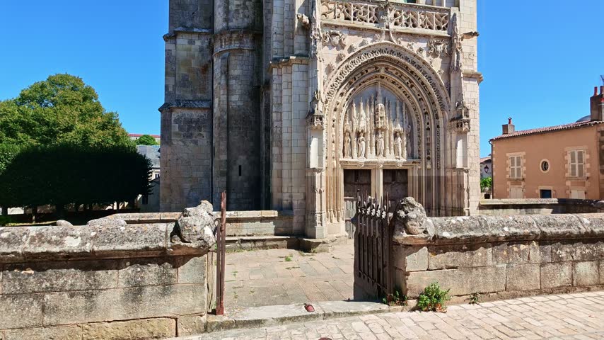 The camera tilts up from the sculpted entrance of Sainte Radegonde Church in Poitiers, France, revealing its stone bell tower and roof against a clear blue sky.