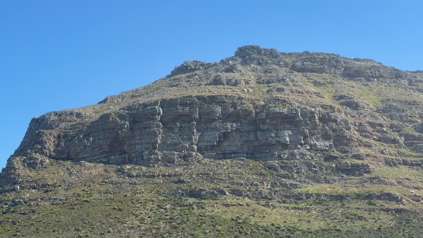 Low-angle view of a rocky mountain face in Cape Town, South Africa, with layered sandstone cliffs and boulders topped by sparse green vegetation. A deep blue cloudless sky above lea