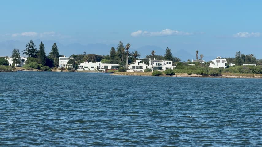 View across the calm, rippled blue water of a lagoon in Muizenberg, Cape Town, South Africa, toward whitewashed waterfront homes among pine and palm trees.