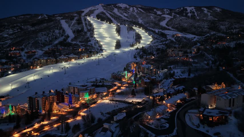 Drone footage resembling Breckenridge, Colorado after dark shows night skiers on bright mountain runs while the snowy resort town glows below with hotels, roads, and holiday lights at night.