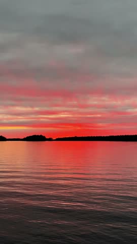 Vivid red sunset reflects on calm lake water with silhouetted tree-lined shore