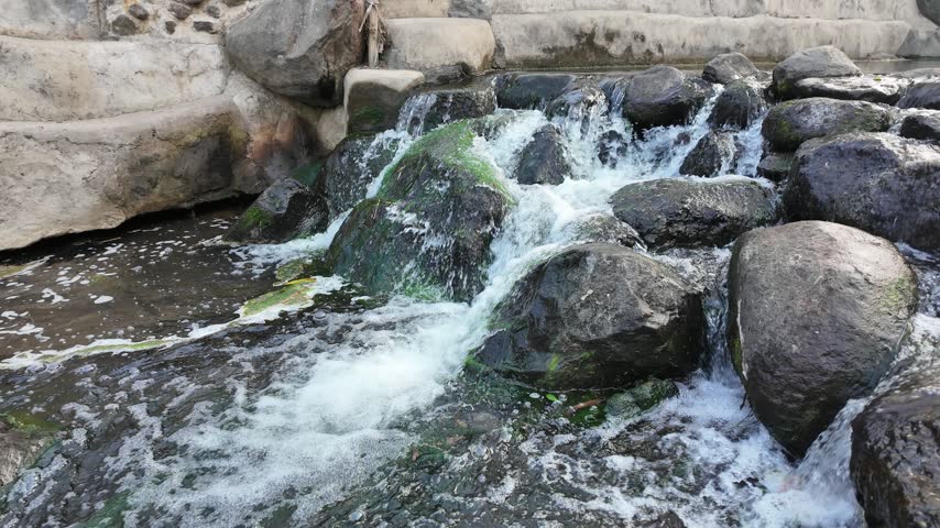 Close-up of clear water cascading over dark mossy rocks in a small rocky river weir, creating white foam and a refreshing natural stream flow.