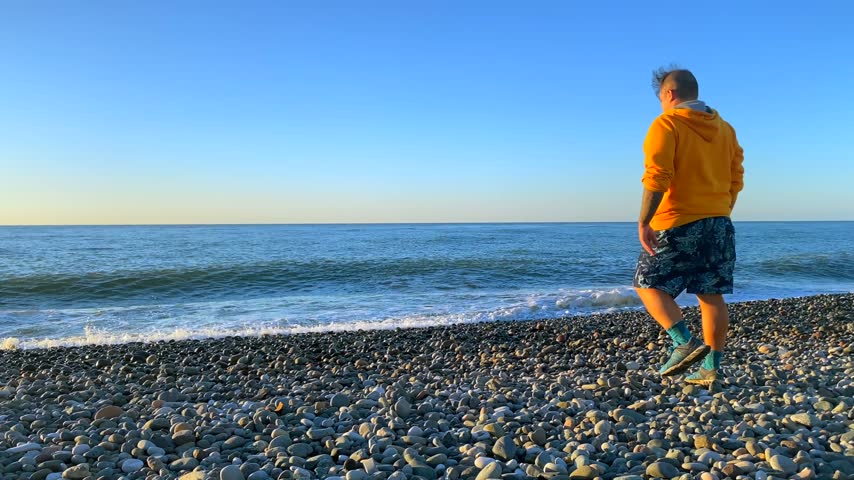 A lone man walks across a stony shoreline toward the Black Sea as small waves reach the beach. The wide coastal view suits travel stories about solo escapes, fresh air and unhurried seaside walks