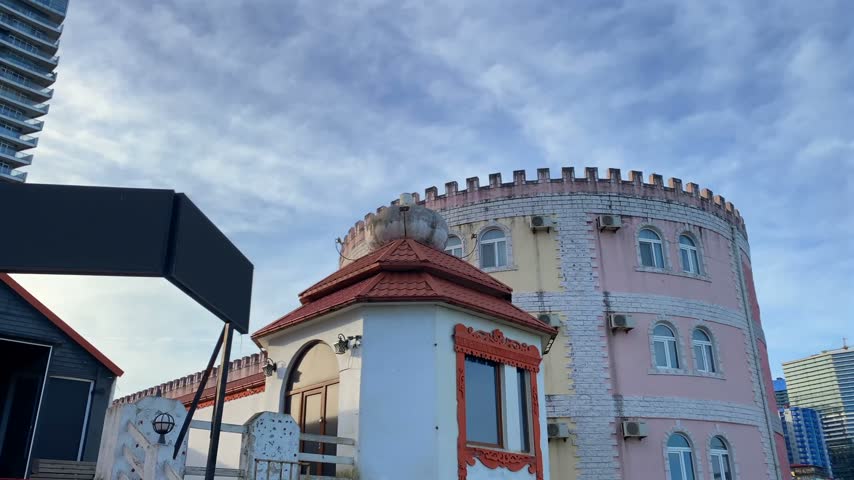 A castle-inspired building with a crenellated roofline and khinkali sculpture stands beside tall high-rise in Batumi. This architectural view suits real estate listings, hospitality and urban tourism