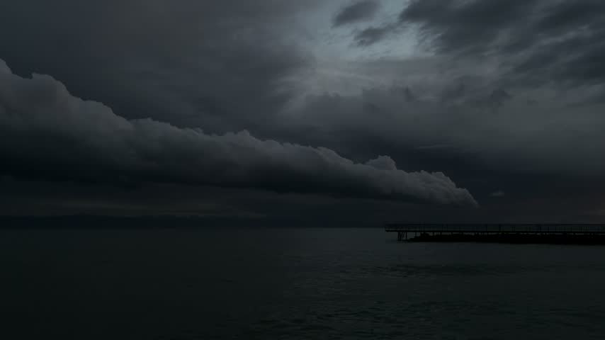 Static shot of ominous roll cloud over dark rippling lake water with silhouetted pier before thunderstorm at dusk, Issyk-Kul, Kyrgyzstan