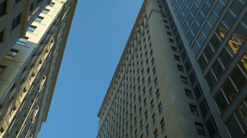 Upward perspective of tall Chicago skyscrapers creating a narrow urban canyon against a clear blue sky. Architectural lines and high-rise structures emphasize dense city development.