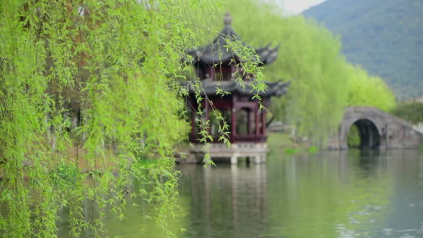 A picturesque traditional Chinese pavilion sits by a calm lake framed by hanging green willow branches and a stone arch bridge in the background.