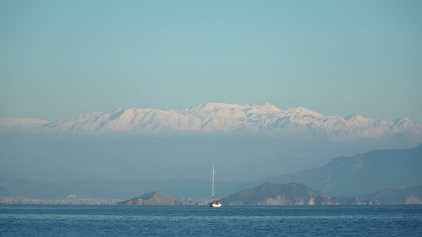 Sailboat on the Mediterranean Sea at Fethiye, Mugla, Turkey, in winter, with snow-capped mountains rising above the haze and the coastline in the distance