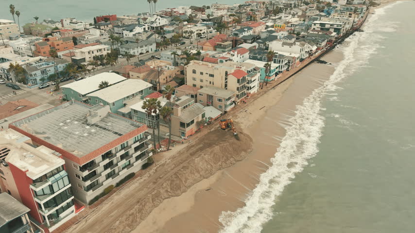 Excavator moves sand beside Long Beach beachfront houses as crews prepare for coastal flooding.