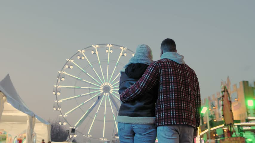 Young couple in love hugging while watching an illuminated ferris wheel at an amusement park