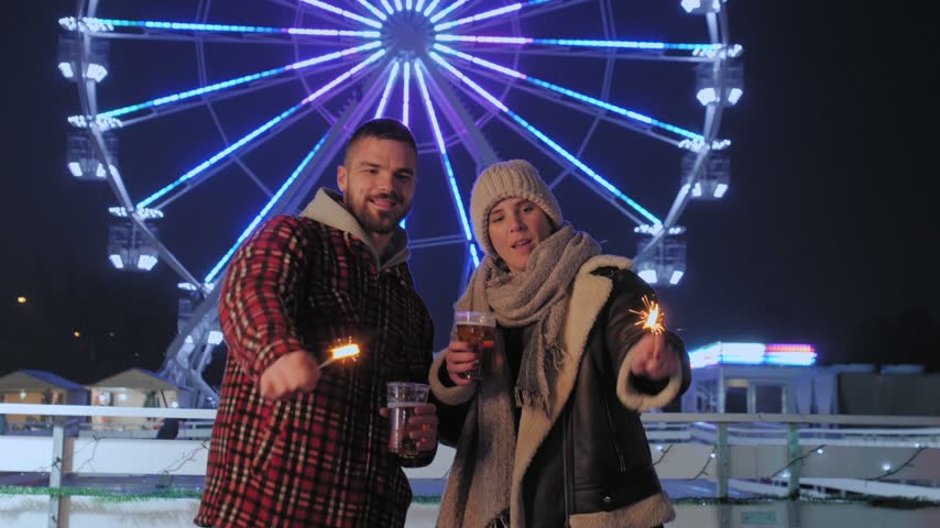 Joyful man and woman having fun with sparklers and drinks at a winter amusement park at night