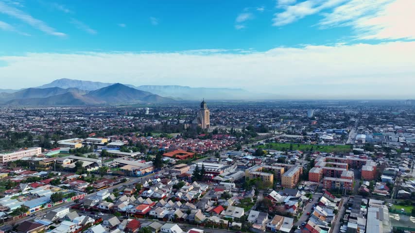 Forward flight toward the Templo Votivo de Maipu, a landmark church tower rising above a vast residential district with apartment blocks beneath hazy mountains. Maipu, Santiago, Chile