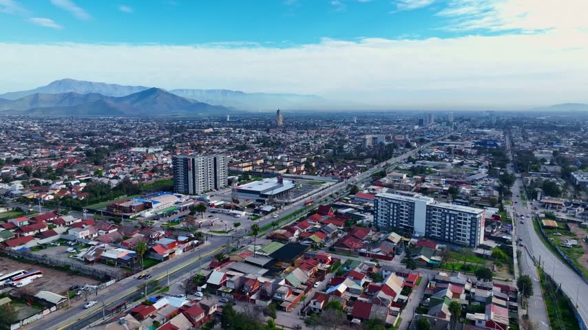 Forward flight over a vast residential district with apartment towers and red tiled rooftops along a main avenue beneath distant hazy mountains. Maipu, Santiago, Chile
