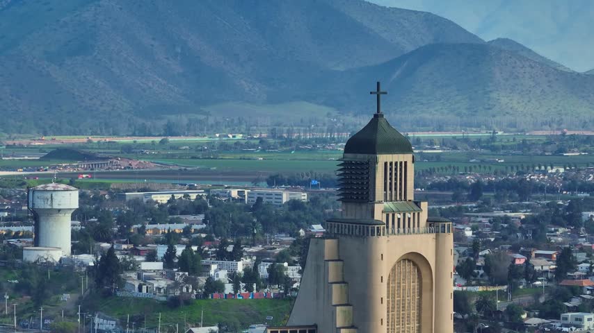 Telephoto panoramic shot of the Templo Votivo de Maipu with its cross-topped tower and arched facade above residential rooftops, a water tower and green farmland beneath hills. Maipu, Santiago, Chile