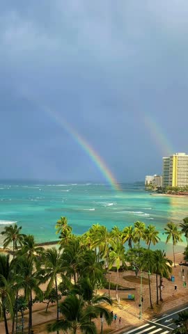 Vibrant rainbow over turquoise ocean water and tropical beach surrounded by lush palm trees