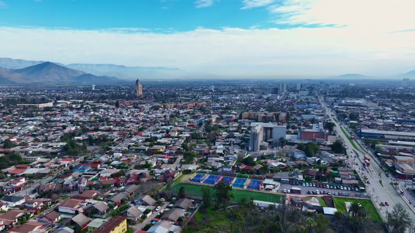 Forward flight over a dense residential district with tennis courts, football pitch and a church tower along a main avenue beneath hazy mountains. Maipu, Santiago, Chile