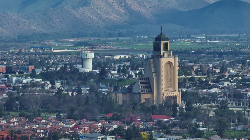Telephoto aerial view of the Templo Votivo de Maipu with its arched facade and cross-topped tower beside a water tower above residential rooftops and green farmland. Maipu, Santiago, Chile