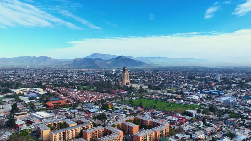 Backward flight revealing the Templo Votivo de Maipu above a vast residential district with apartment blocks and a wide green park beneath layered mountains. Maipu, Santiago, Chile