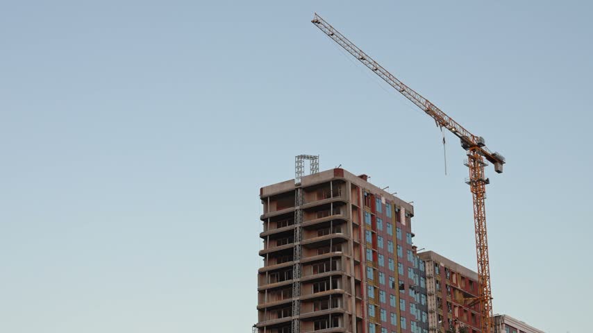A towering yellow crane stands tall against a pale sky, slowly pivoting its long arm above the structure of a multi-story building that remains under active development in the city during the daytime.