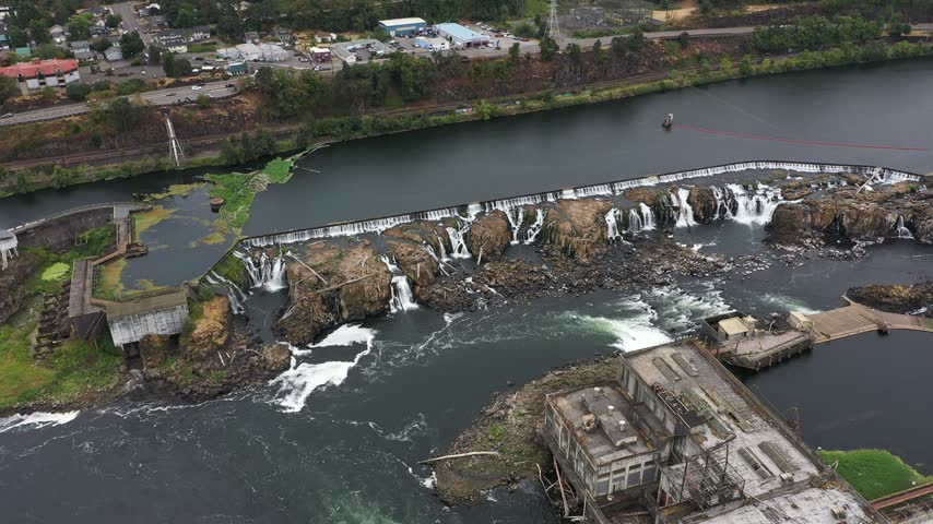 4K aerial drone footage of Willamette Falls on the Willamette River between Oregon City and West Linn, near Portland, Oregon, USA. Scenic aerial view showcases the historic   Hydroelectric Plant, former paper mill buildings, industrial facilities, electrical substations, power generation infrastructure and cascading waterfalls flowing over rocky river formations. The surrounding landscape includes forested hills, residential neighborhoods, roads, bridges and urban areas of Oregon City and West Linn in the Portland metropolitan area. This historic industrial complex represents the region