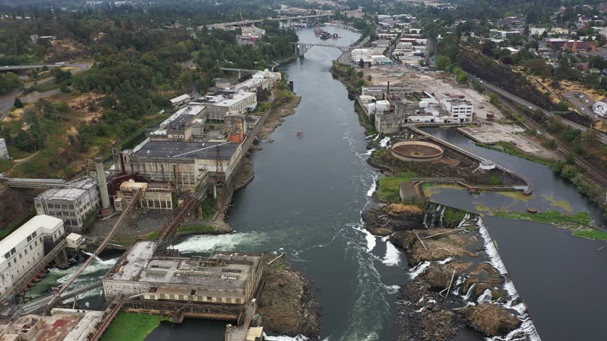 4K aerial drone footage of Willamette Falls on the Willamette River between Oregon City and West Linn, near Portland, Oregon, USA. Scenic aerial view showcases the historic   Hydroelectric Plant, former paper mill buildings, industrial facilities, electrical substations, power generation infrastructure and cascading waterfalls flowing over rocky river formations. The surrounding landscape includes forested hills, residential neighborhoods, roads, bridges and urban areas of Oregon City and West Linn in the Portland metropolitan area. This historic industrial complex represents the region