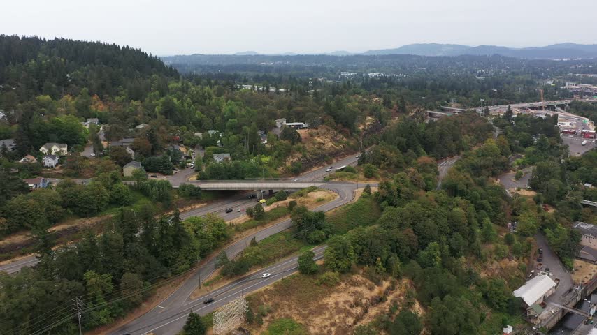 4K aerial drone footage of Oregon City near Portland, Oregon, USA, showing Highway 99E winding through forested hills and residential neighborhoods along the Willamette River on a gloomy, overcast day. Elevated views capture an electrical power substation with transformers, transmission towers and power lines, alongside busy roads, highway intersections, bridges, residential homes and surrounding urban development. The landscape features dense evergreen trees, wooded hillsides, industrial and commercial buildings, and transportation infrastructure near the river. Scenic Pacific Northwest footage useful for electrical grid infrastructure, electricity distribution, power transmission, highway traffic, suburban development, transportation, urban planning, Oregon geography and environmental B-roll.
