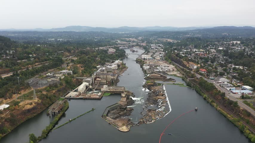 4K aerial drone footage of Willamette Falls on the Willamette River between Oregon City and West Linn, near Portland, Oregon, USA. Scenic aerial view showcases the historic   Hydroelectric Plant, former paper mill buildings, industrial facilities, electrical substations, power generation infrastructure and cascading waterfalls flowing over rocky river formations. The surrounding landscape includes forested hills, residential neighborhoods, roads, bridges and urban areas of Oregon City and West Linn in the Portland metropolitan area. This historic industrial complex represents the region
