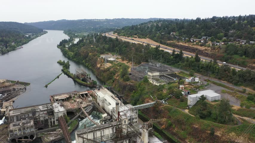 4K aerial drone footage of Willamette Falls on the Willamette River between Oregon City and West Linn, near Portland, Oregon, USA. Scenic aerial view showcases the historic   Hydroelectric Plant, former paper mill buildings, industrial facilities, electrical substations, power generation infrastructure and cascading waterfalls flowing over rocky river formations. The surrounding landscape includes forested hills, residential neighborhoods, roads, bridges and urban areas of Oregon City and West Linn in the Portland metropolitan area. This historic industrial complex represents the region