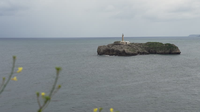 Rocky Mouro Island with its stone lighthouse in a calm grey sea, framed by blurred yellow wildflowers in the foreground on a cloudy day at the entrance of Santander Bay, Spain